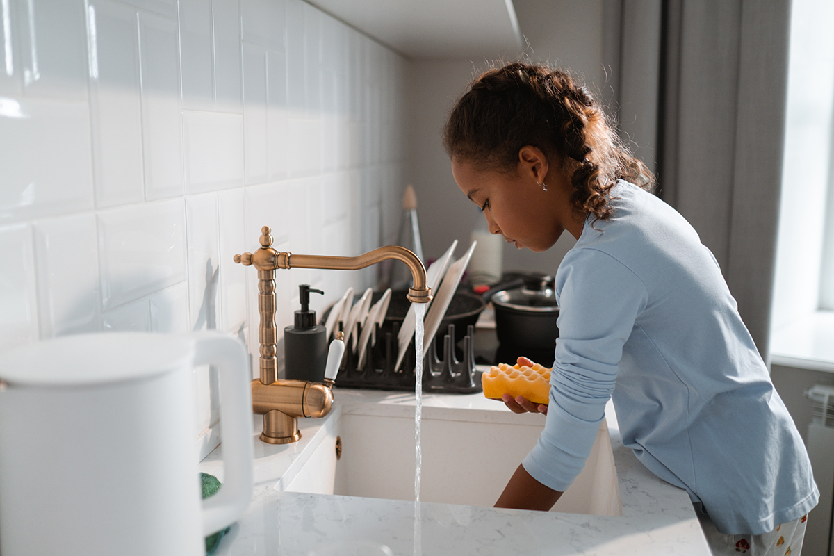 a young girl washing dishes in a sink with a gold faucet.