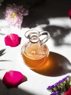 a glass pitcher of golden honey syrup next to rose petals.