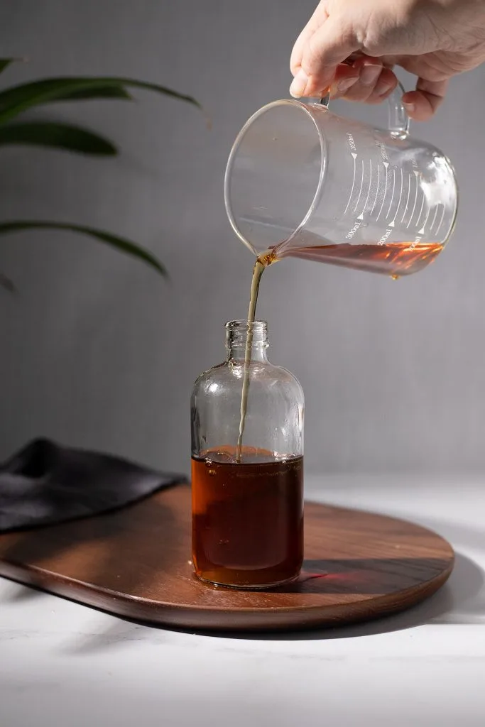 pouring dark brown demerara syrup into an apothecary bottle.