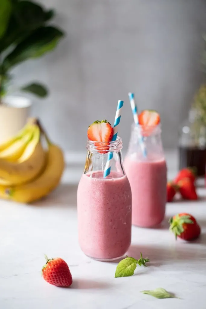 two glass bottles full of pink smoothies garnished with strawberries and blue paper straws.