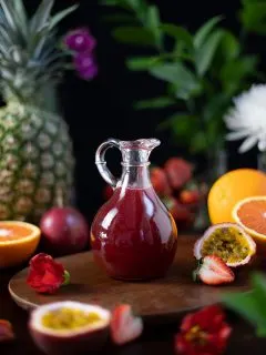a glass pitcher of dark red syrup next to tropical fruits.