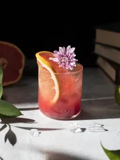 a rocks glass filled with a pink drink garnished with a grapefruit slice and a purple flower.