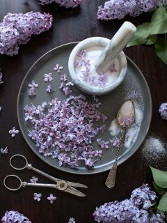 a mortar and pestle with white sugar surrounded by lilac flowers.