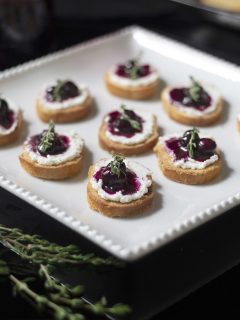 a white platter of small crostinis topped with blueberry compote and thyme.