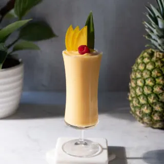 a bright yellow smoothie in a tall hurricane glass garnished with fruit.