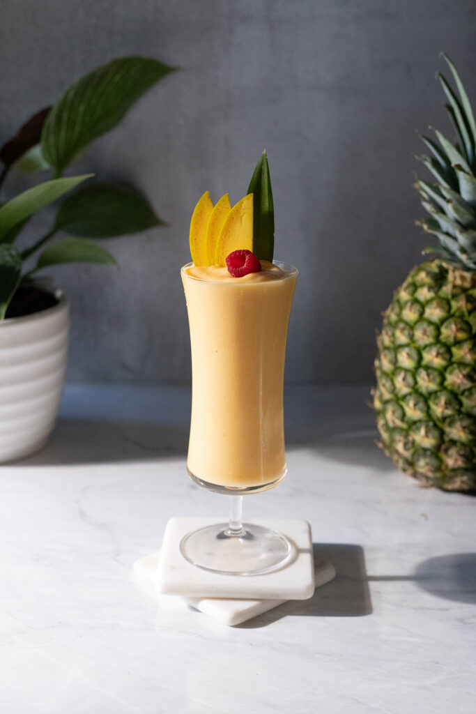 a bright yellow smoothie in a tall hurricane glass garnished with fruit.