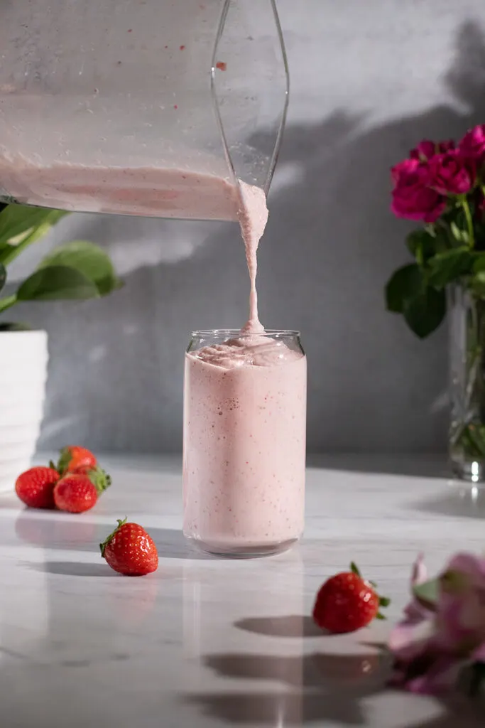 pouring a creamy pale pink smoothie into a tall glass.