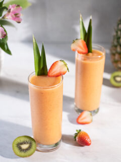 two orange smoothies in tall glasses garnished with pineapple leaves.