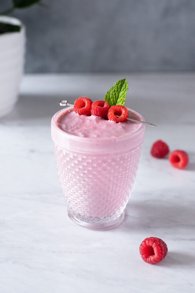 a pink smoothie in a fancy glass with three skewered raspberries.
