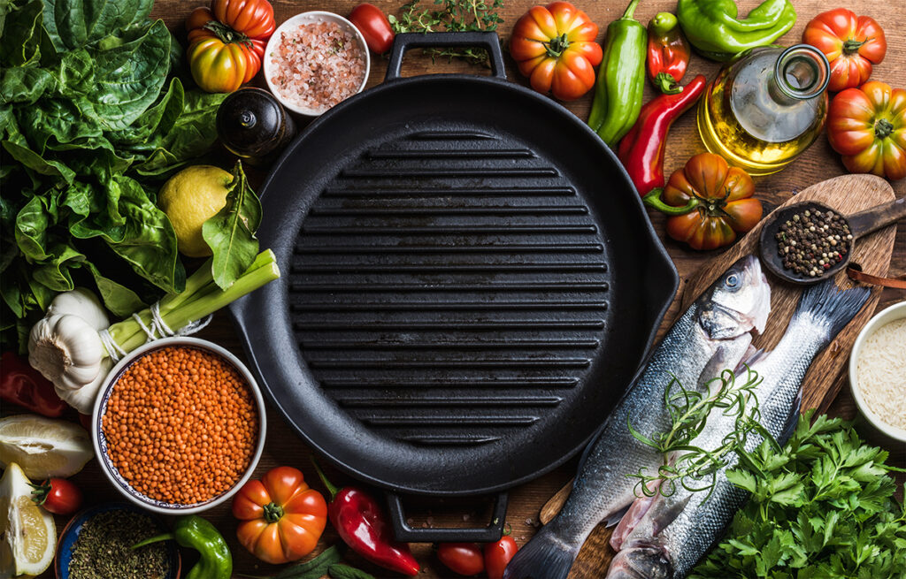 a cast iron grill pan surrounded by fresh produce, fish, and spices.