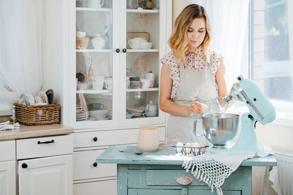 a blond woman cracking an egg into a pale blue stand mixer in a sunny kitchen.