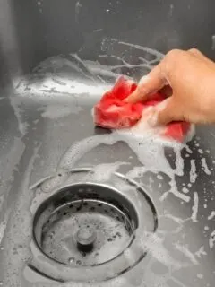 a hand scrubbing a stainless steel sink with a soapy red sponge.